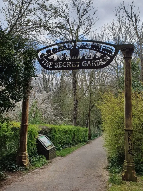 Eisenbogen mit 'The Secret Garden'-Schild über einem Gartenweg, umgeben von Bäumen und Sträuchern.