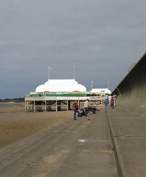 People walking on a concrete promenade towards a pier with a white pavilion.