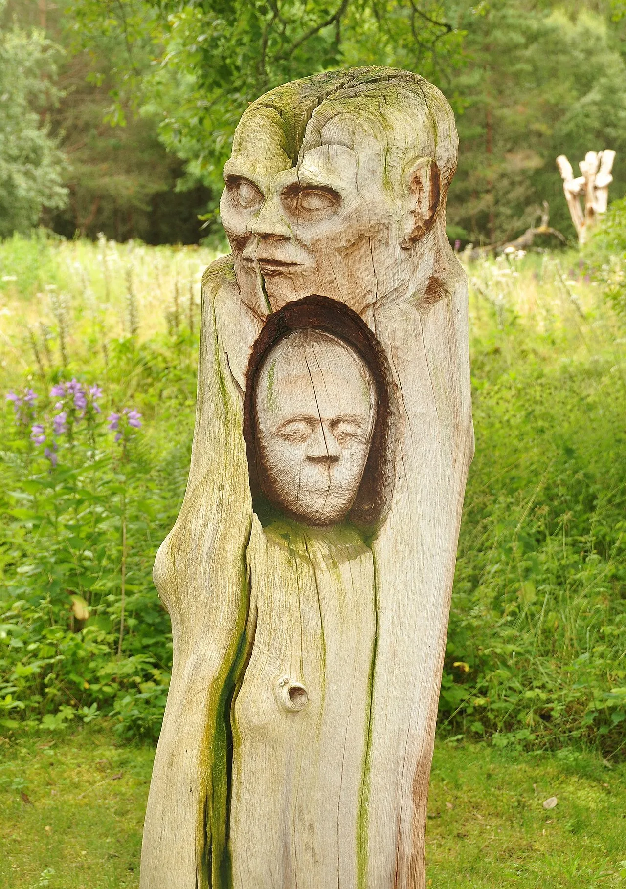 Holzskulptur mit zwei Gesichtern, eines größer oben, eines kleiner unten, im Park.