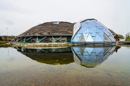 A modern leisure centre with a glass dome and thatched roof reflected in water.
