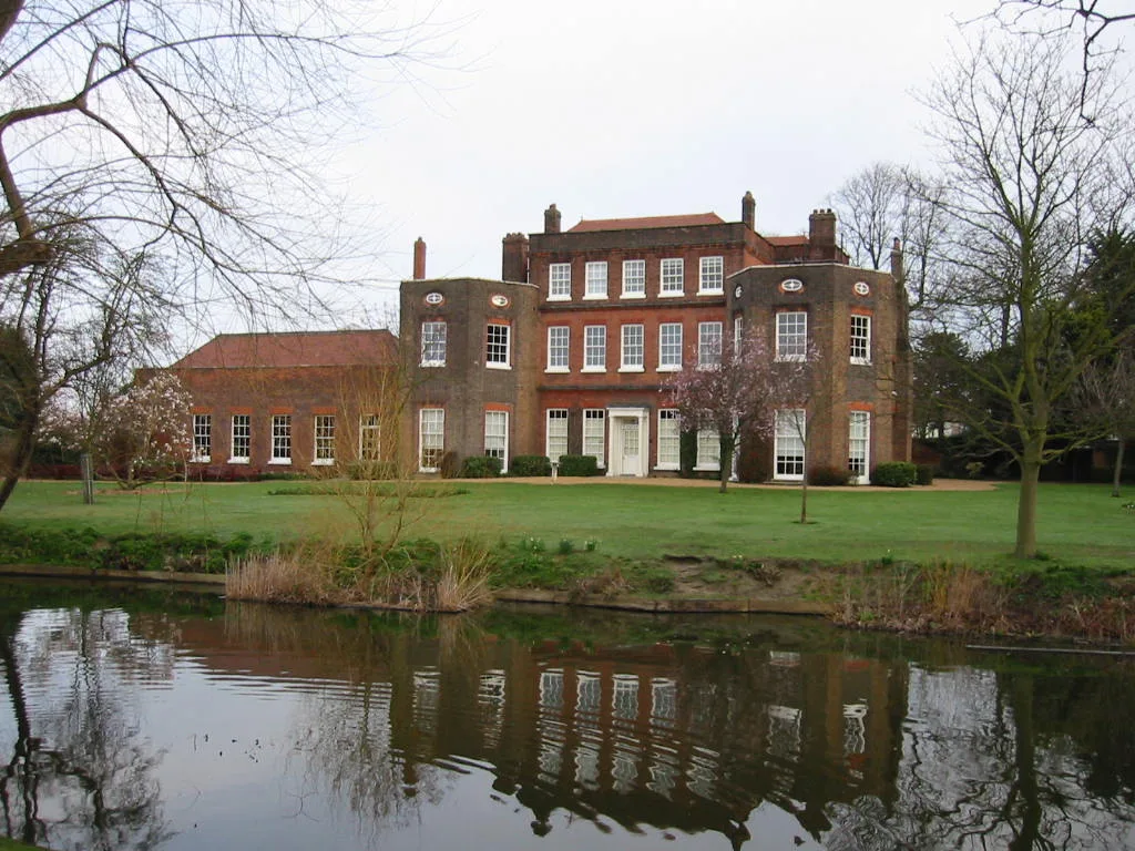 A red brick house with multiple windows reflected in a pond with trees around.