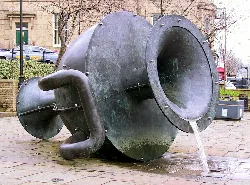 Große Metallskulptur eines geneigten Teekannen mit Wasser, das aus dem Ausguss fließt.