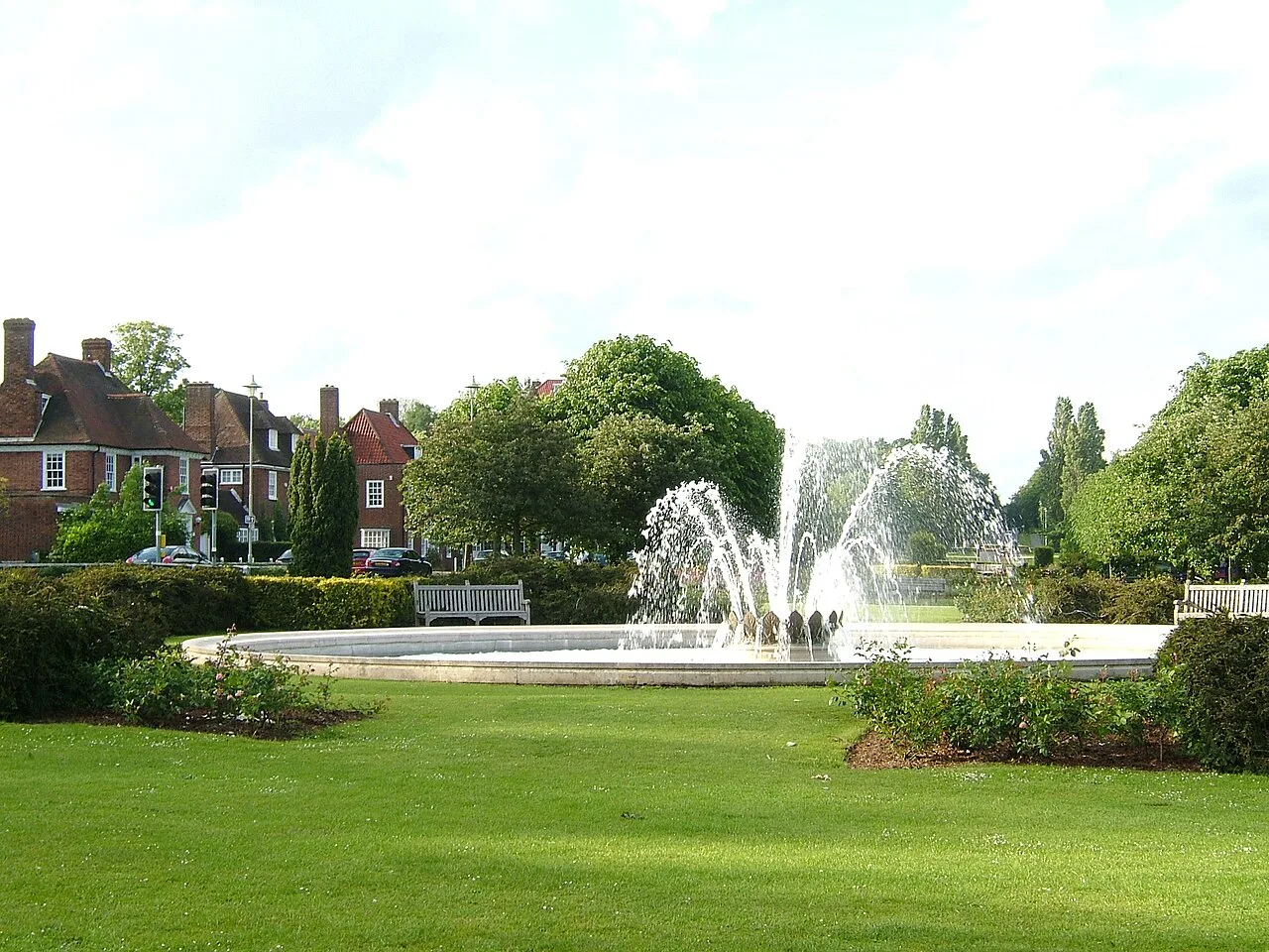 Fuente con chorros de agua en un parque rodeado de árboles y casas.