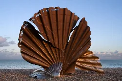 A large metallic scallop shell sculpture on a beach with sea and sky in the background.