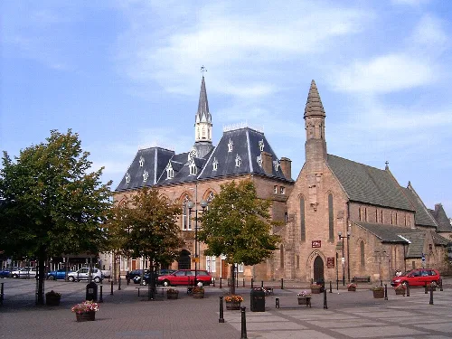Rathaus und Kirchengebäude mit Bäumen und parkenden Autos davor.