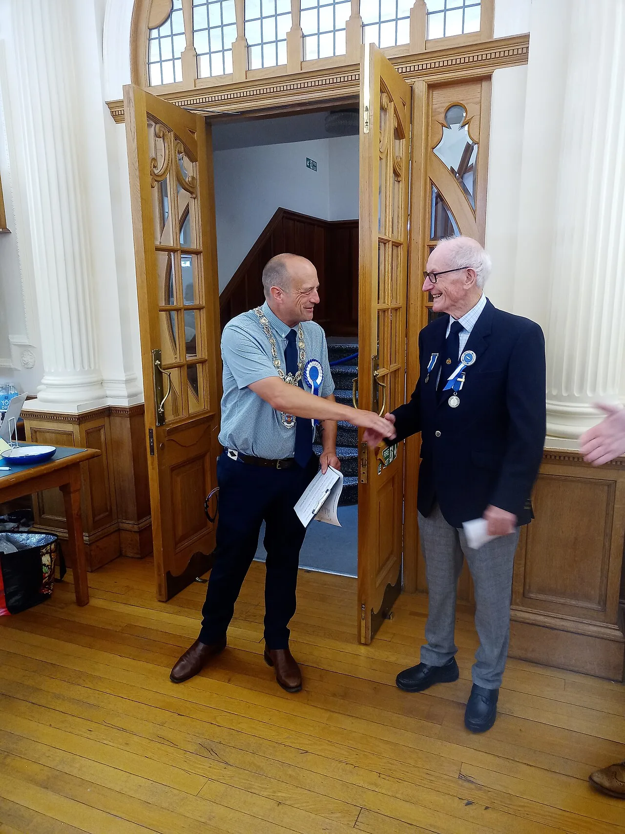Two men shaking hands inside Kelso town hall, one in a suit and the other in a casual shirt.