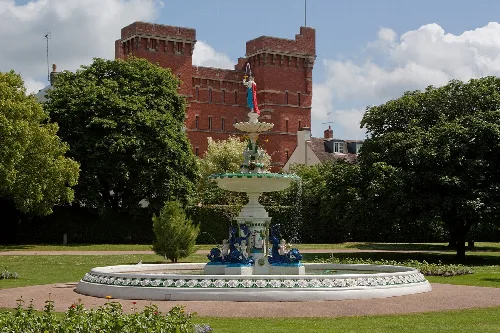 Colorful fountain with a statue on top in a park, with a red brick building behind.