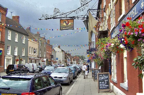 Blick auf die Hauptstraße in Stony Stratford mit parkenden Autos, Geschäften und Blumenampeln.
