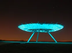 Eine große, kreisförmige, blau beleuchtete Struktur auf drei Beinen vor dunklem Himmel.