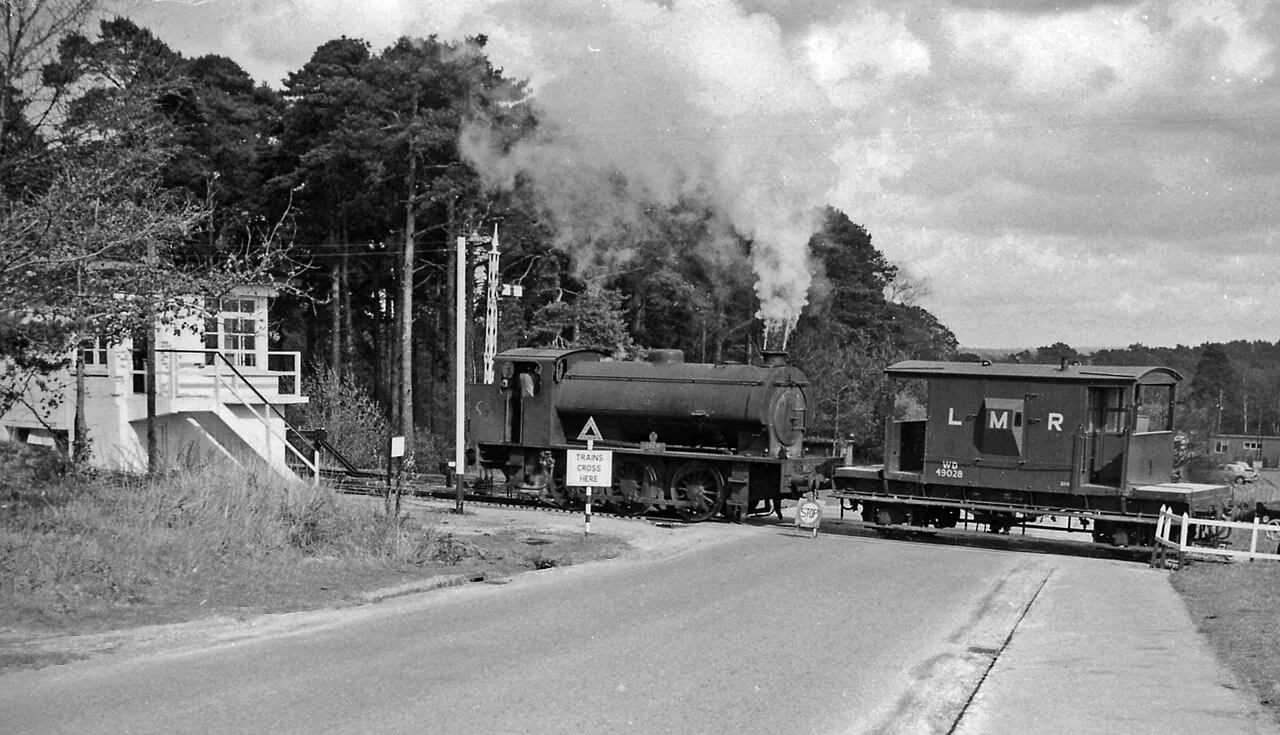 Longmoor Military Railway crossing a road at Bordon, 1963.
View eastwards near Bordon, probably on the B2131 road - [but I am not quite certain exactly where, as the network of the LMR was complex and has now completely disappeared]. There is a signalbox (freshly painted in Army fashion) on the left, but no crossing-gates. Crossing with a LMR brakevan (British Railways standard 20-ton design, numbered WD 49028) is a typical WD Austerity 0-6-0ST, WD 102 'CAEN'. This is in the latter days of the LMR, as it was all closed by October 1969.