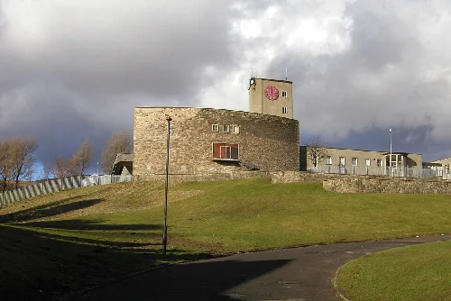 Ein steinernes Schulgebäude auf einem grasbewachsenen Hügel mit Uhrturm und wolkigem Himmel.