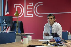 Premierminister Rishi Sunak an einem Schreibtisch mit Laptop und Tassen vor einem roten DEC-Schild.
