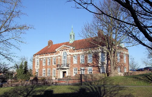 A red brick building with a central clock tower and multiple windows, surrounded by trees and a black fence.