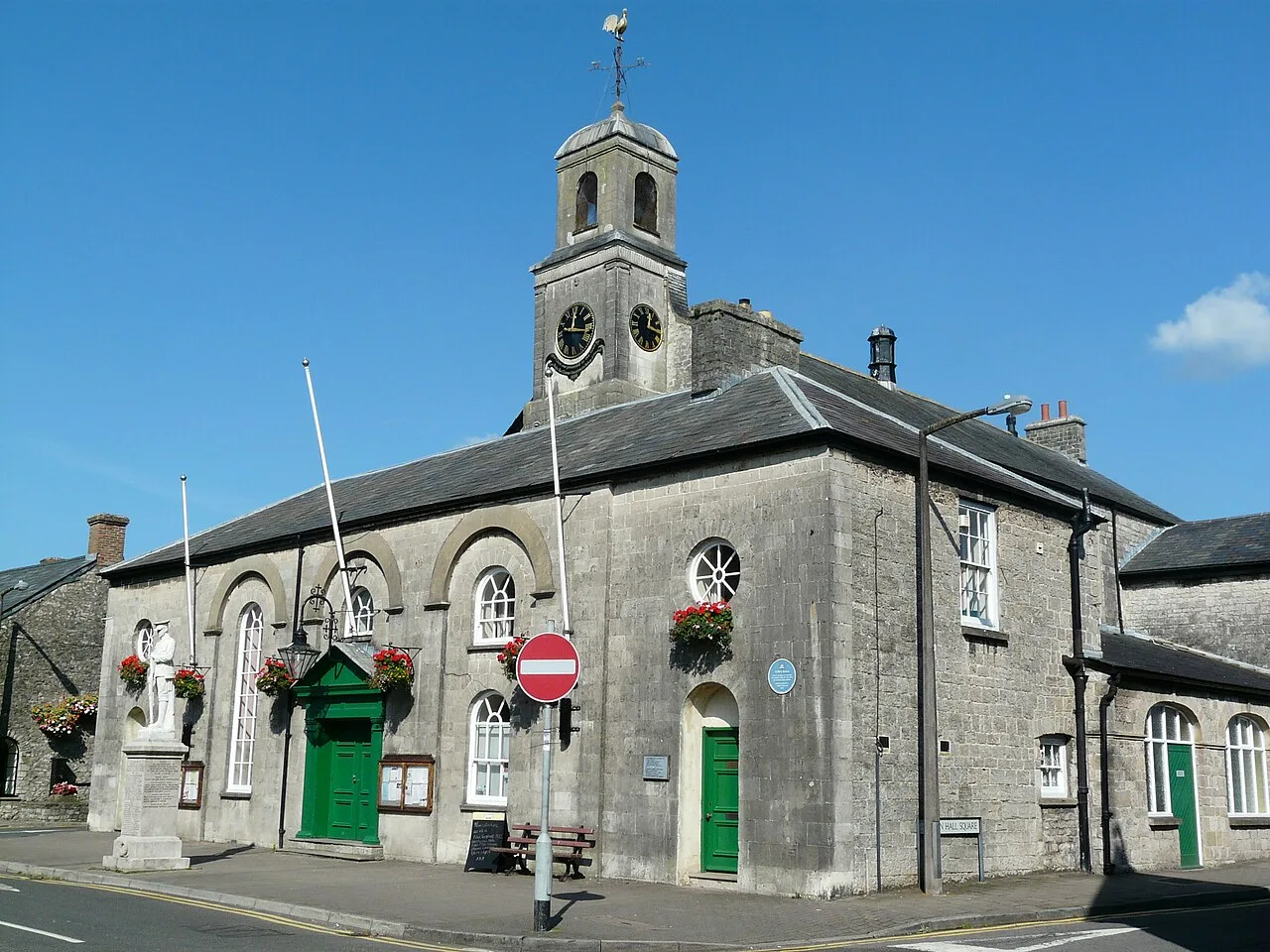 Edificio de piedra con puertas verdes, torre del reloj y cestas de flores en Cowbridge.