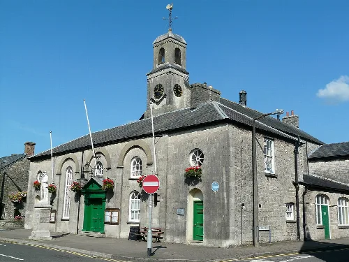 Edificio de piedra con puertas verdes, torre del reloj y cestas de flores en Cowbridge.
