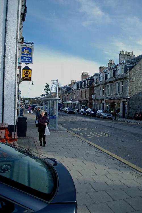 View of Banchory high street with buildings, cars, and a woman walking.