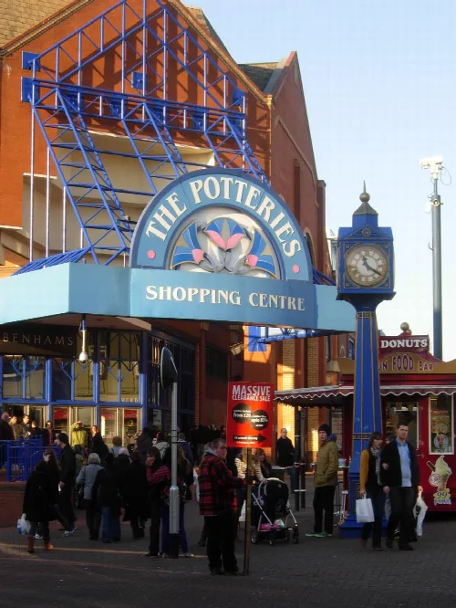 Entrance to The Potteries Shopping Centre with a blue clock and people outside.