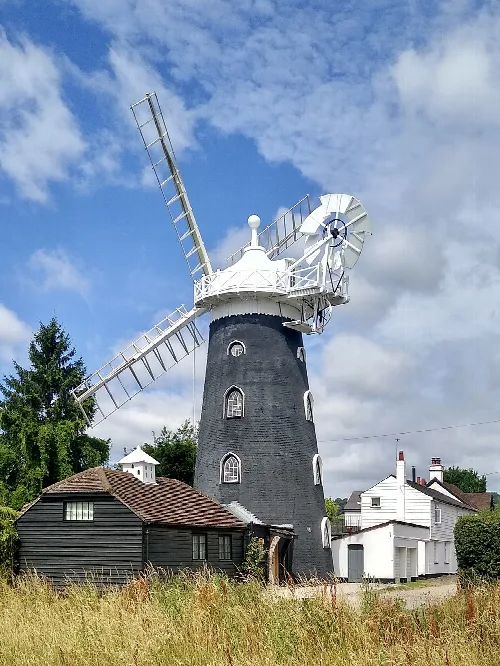 A tall, black windmill with white sails on Batts Hill, surrounded by small buildings and greenery.