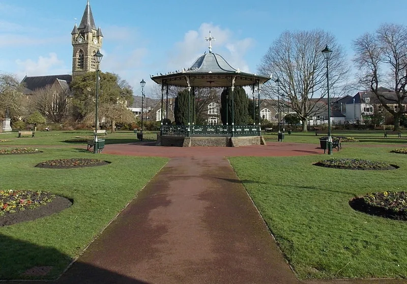 Quiosco octagonal en Victoria Gardens, Neath, con una torre de iglesia al fondo.