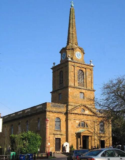 Steinkirche mit hohem Turm und Uhr, umgeben von Bäumen und parkenden Autos.